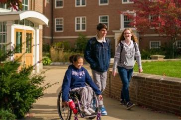 Three UNH students, one in a wheelchair and two standing alongside her, walk and wheel in the courtyard at UNH's Murkland Hall.